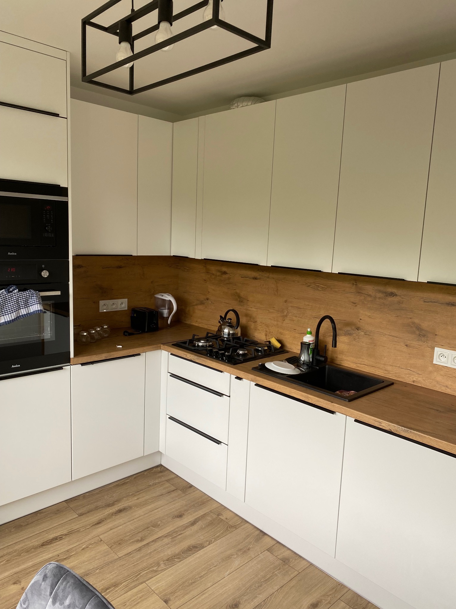 Nowoczesna kuchnia w bieli i drewnie: białe szafki z czarnymi uchwytami, drewniany blat i panel nad blatem, czarny zlew i bateria, płyta gazowa z czajnikiem, oświetlenie sufitowe w stylu...