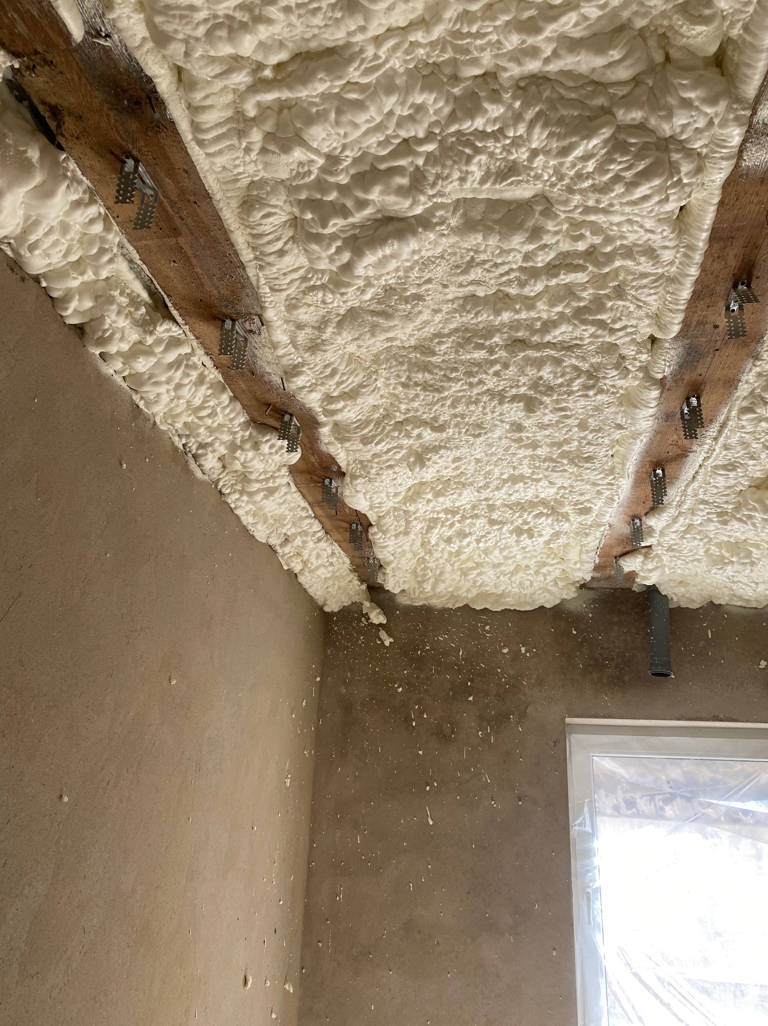 Ocieplenie poddasza pianką PUR, widoczne drewniane belki stropowe i metalowe uchwyty. Ściany surowe, okno zabezpieczone folią.