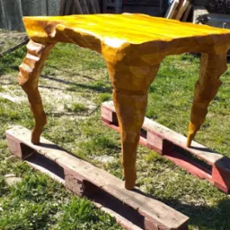 Stół z litego drewna o nieregularnym kształcie blatu i rzeźbionych nogach, ustawiony na dwóch drewnianych paletach na trawie.