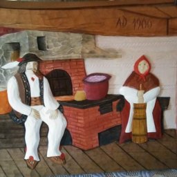 Klimkówka - Drewniana płaskorzeźba przedstawiająca scenę z życia góralskiej rodziny, z datą AD 1900, ukazująca mężczyznę siedzącego obok pieca i kobietę ubijającą masło w drewnianej beczce.