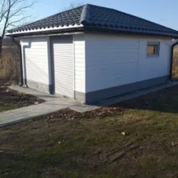 Mały, biały budynek gospodarczy z szarym dachem i rolowanymi drzwiami, otoczony trawnikiem i ścieżką z kostki.