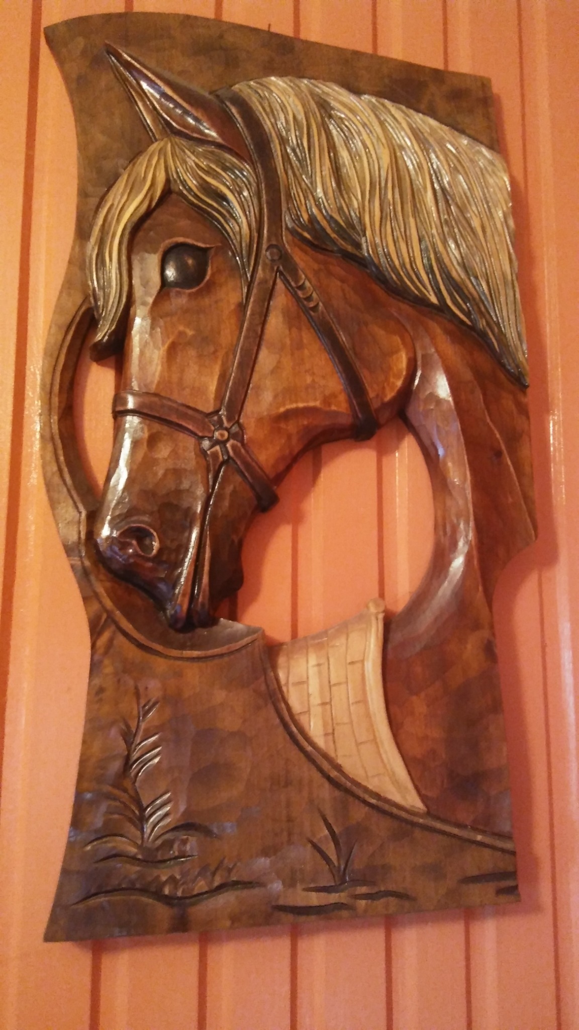 Ręcznie rzeźbiona w drewnie płaskorzeźba przedstawiająca głowę konia w uprzęży, z widocznymi detalami sierści i roślinnym motywem u dołu, zawieszona na ścianie z pionowymi panelami.