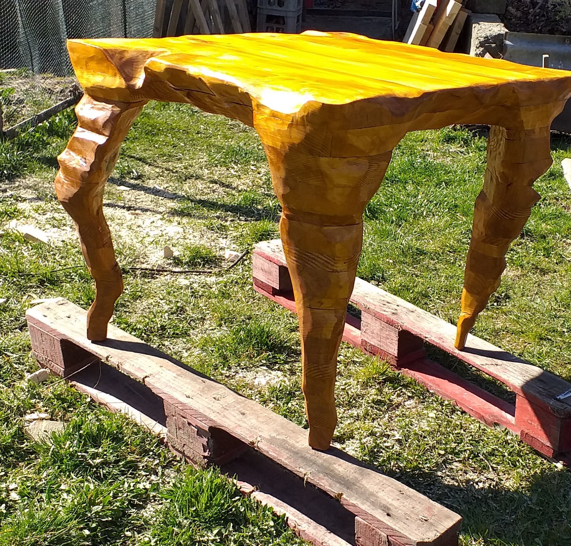 Stół z litego drewna o nieregularnym kształcie blatu i rzeźbionych nogach, ustawiony na dwóch drewnianych paletach na trawie.