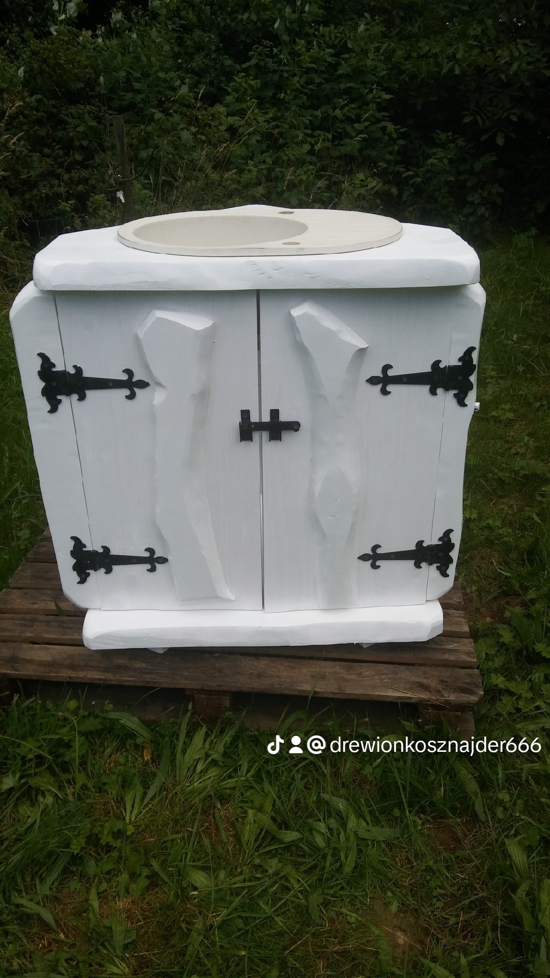 Biała, stylizowana szafka z drewnianymi drzwiczkami, ozdobiona czarnymi zawiasami i zamkiem, z umywalką na górze, umieszczona na drewnianej palecie na trawie.