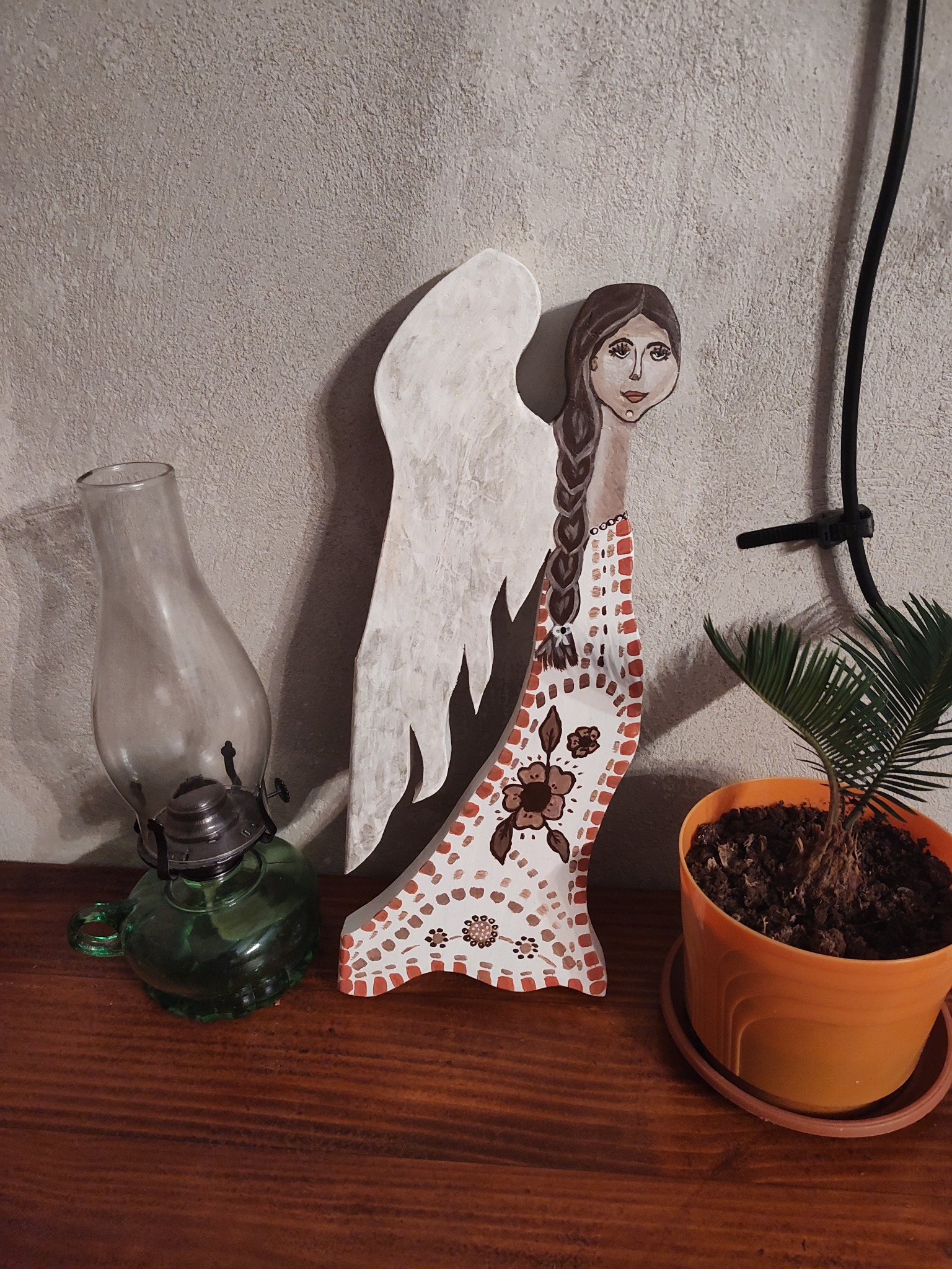 Ręcznie malowana drewniana figurka anioła z ludowym wzorem, obok zielona lampa naftowa i mała palma w pomarańczowej doniczce na drewnianym blacie.