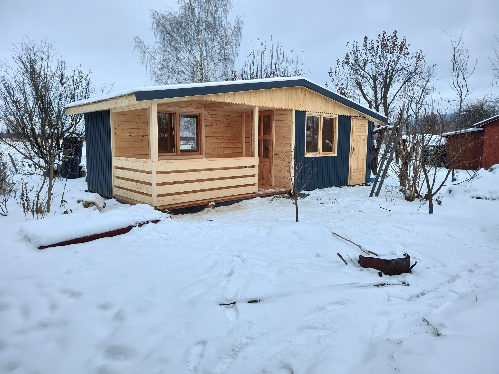 Drewniana konstrukcja z niebieskimi panelami w zimowym krajobrazie, z widocznymi oknami i drzwiami, otoczona śniegiem i gołymi drzewami.