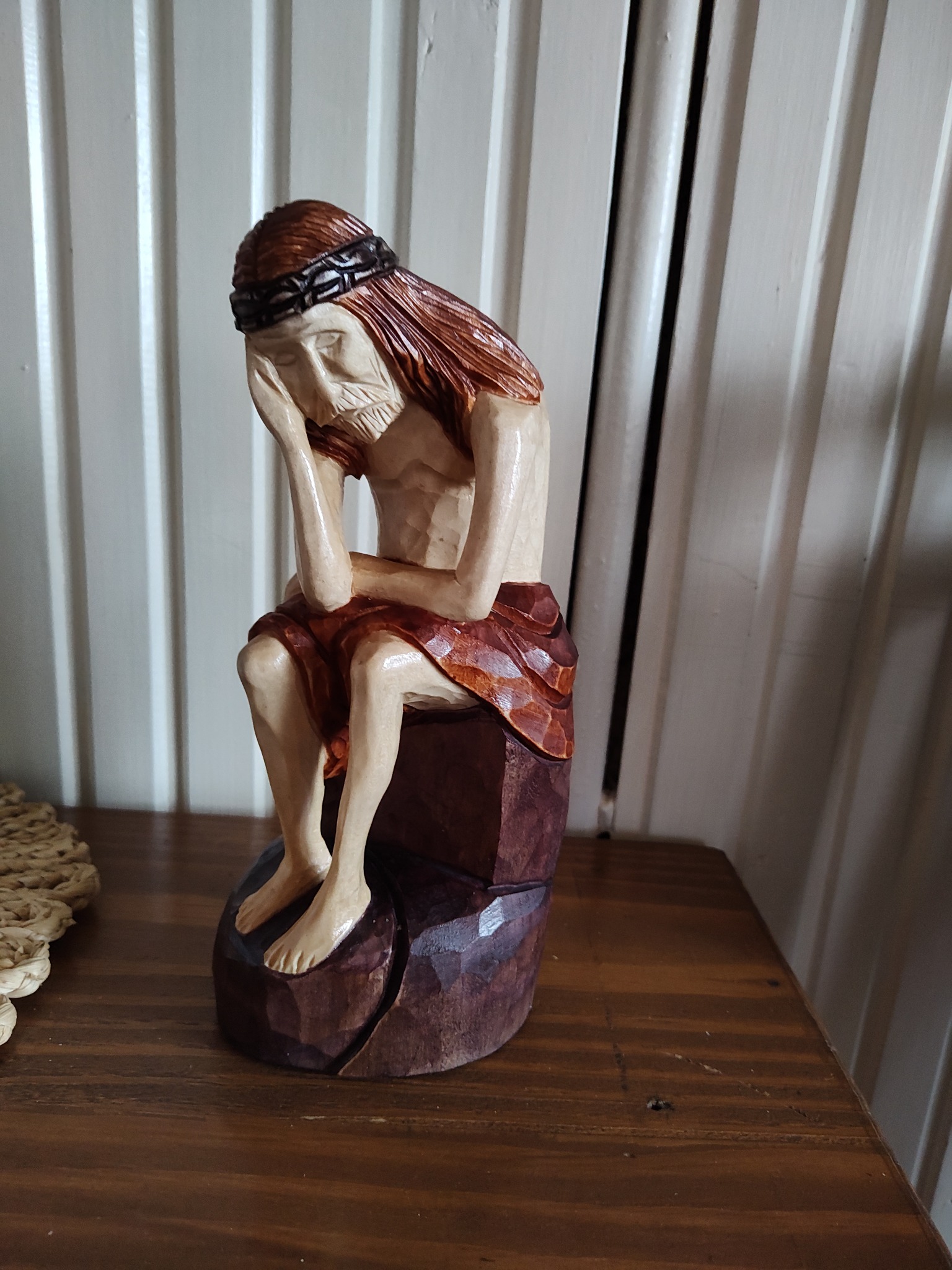 Ręcznie rzeźbiona, drewniana figurka przedstawiająca siedzącego Jezusa z koroną cierniową, umieszczona na drewnianym stoliku z widoczną fakturą drewna i tłem z białych paneli.