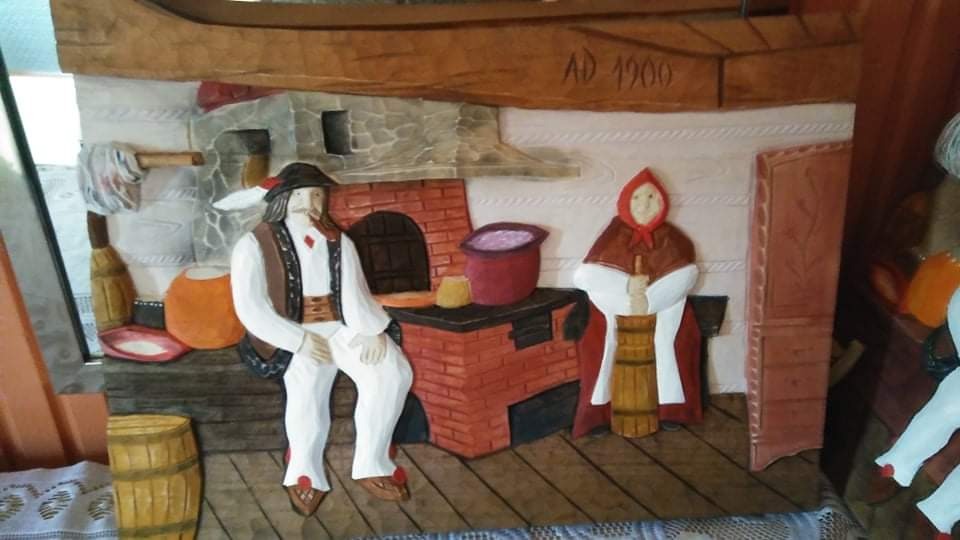 Drewniana płaskorzeźba przedstawiająca scenę z życia góralskiej rodziny, z datą AD 1900, ukazująca mężczyznę siedzącego obok pieca i kobietę ubijającą masło w drewnianej beczce.