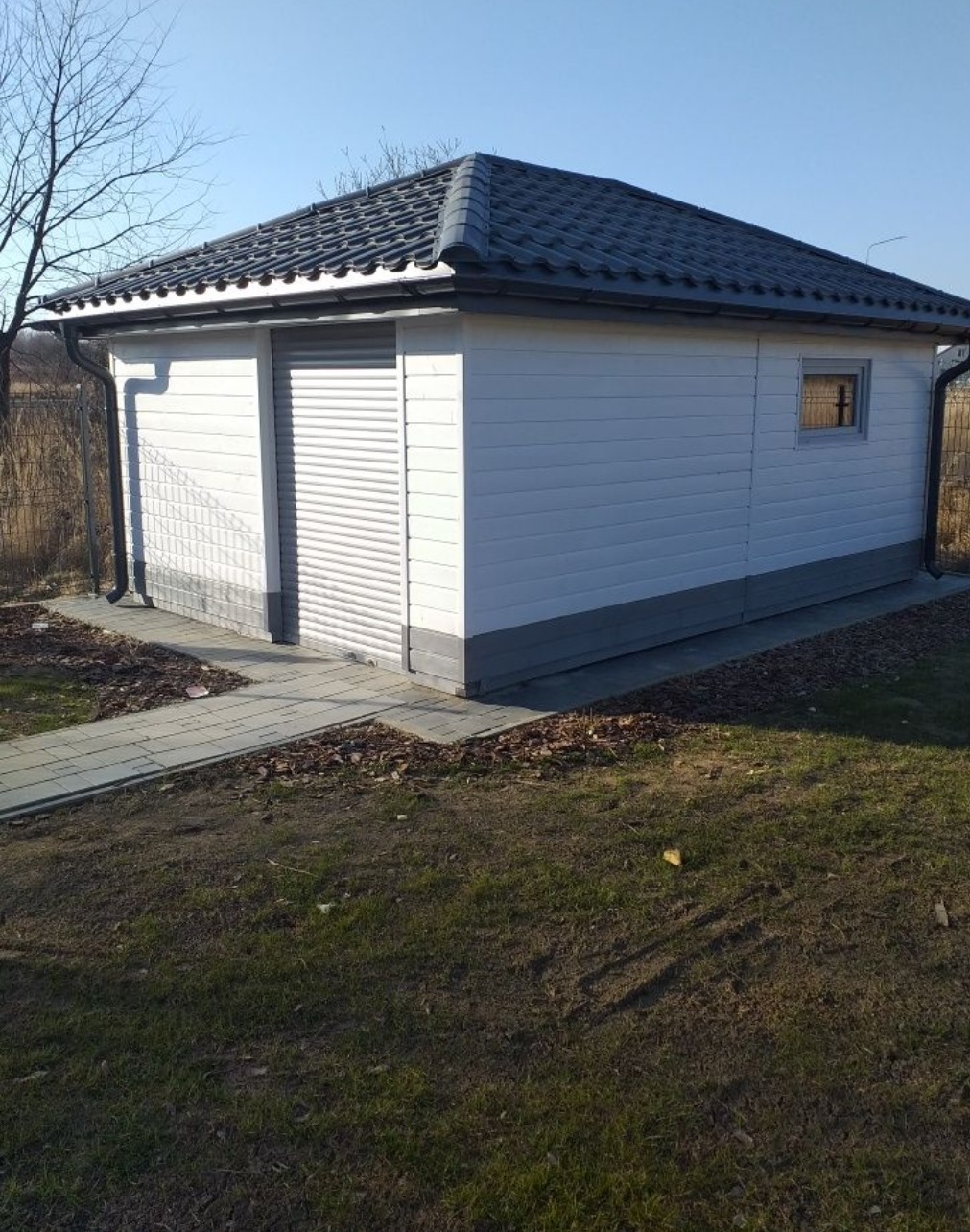 Mały, biały budynek gospodarczy z szarym dachem i rolowanymi drzwiami, otoczony trawnikiem i ścieżką z kostki.