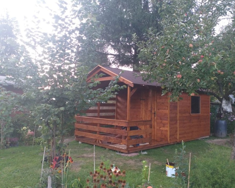 Drewniana altana z zadaszonym tarasem, pomalowana na brązowo, otoczona zielenią ogrodu z drzewami owocowymi i kwiatami, widoczna puszka z farbą.