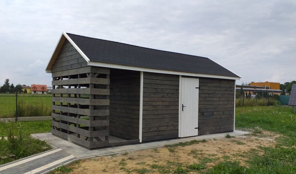 Drewniana konstrukcja z dachem krytym papą, z jedną ścianą ażurową i białą, prostą furtką, na obrzeżu trawnika