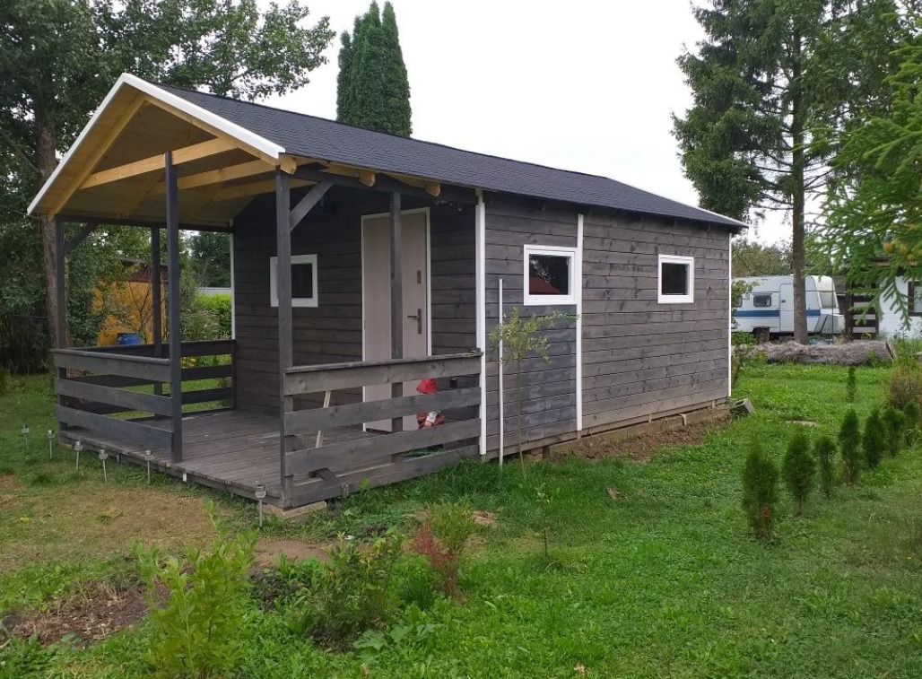 Drewniany domek letniskowy z zadaszonym tarasem, pomalowany na szaro, z czarnym dachem, na tle zielonego trawnika i drzew.