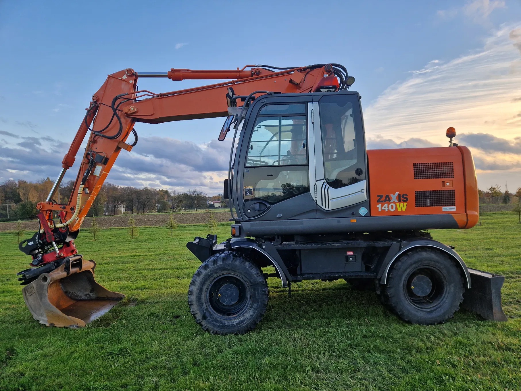 Pomarańczowa koparka kołowa Zaxis 140W stoi na zielonej trawie z łyżką skierowaną w dół, na tle pola i zachodzącego słońca.