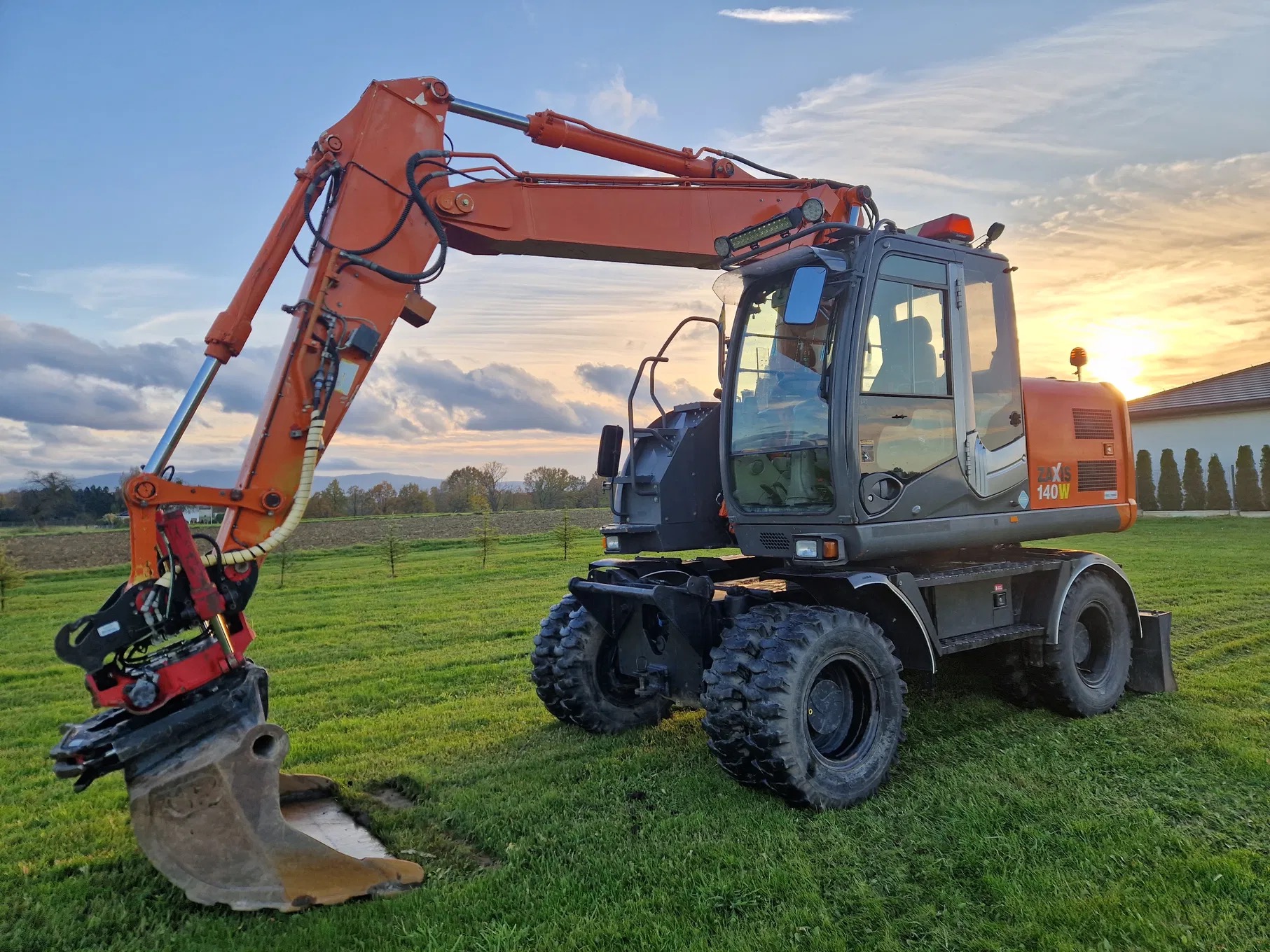 Pomarańczowo-szara koparka kołowa Hitachi ZX140W na zielonej trawie, w tle zachodzące słońce i częściowo widoczny budynek.