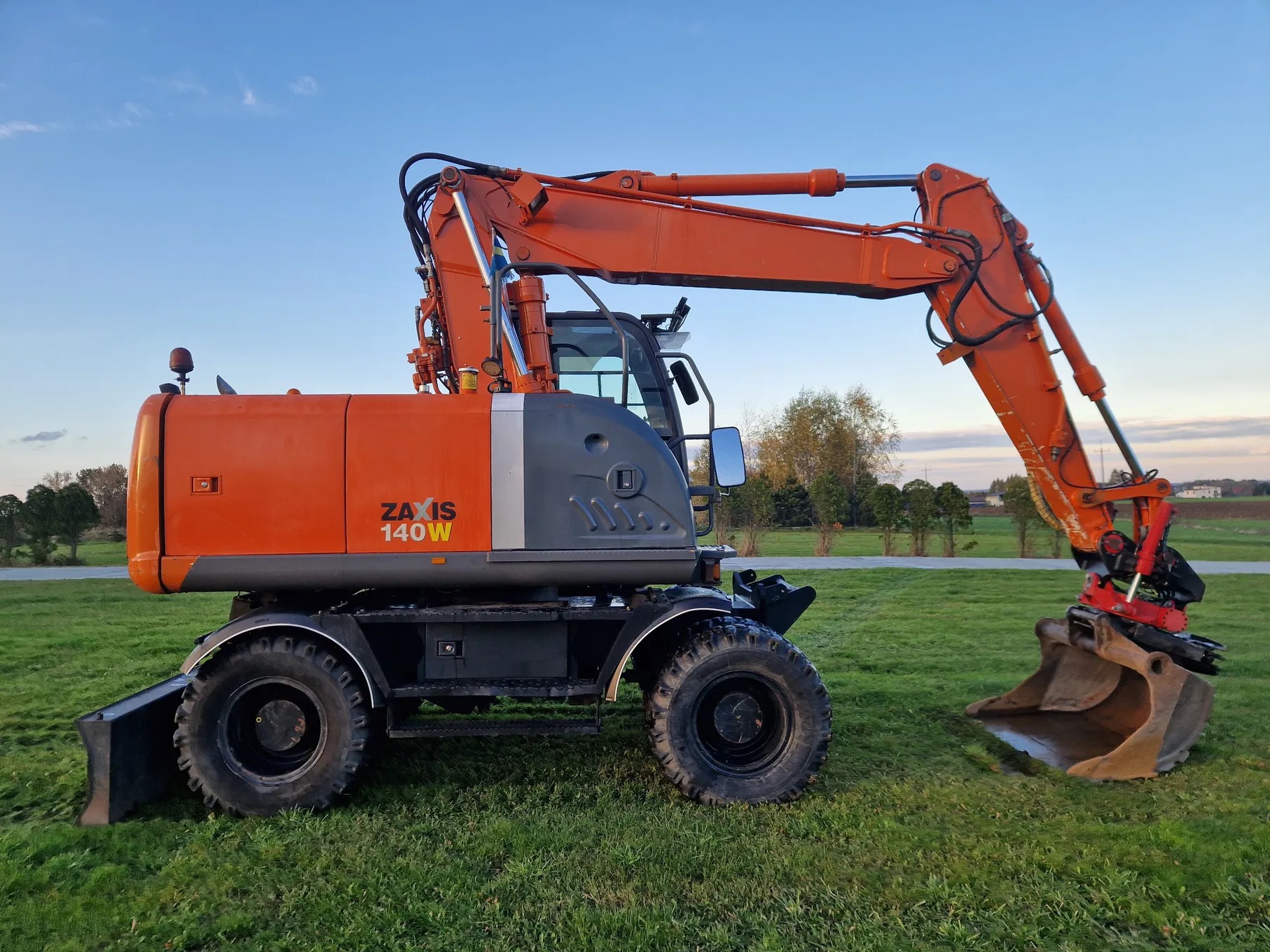 Pomarańczowa koparka kołowa Zaxis 140W z łyżką, stojąca na trawie pod błękitnym niebem o zmierzchu.