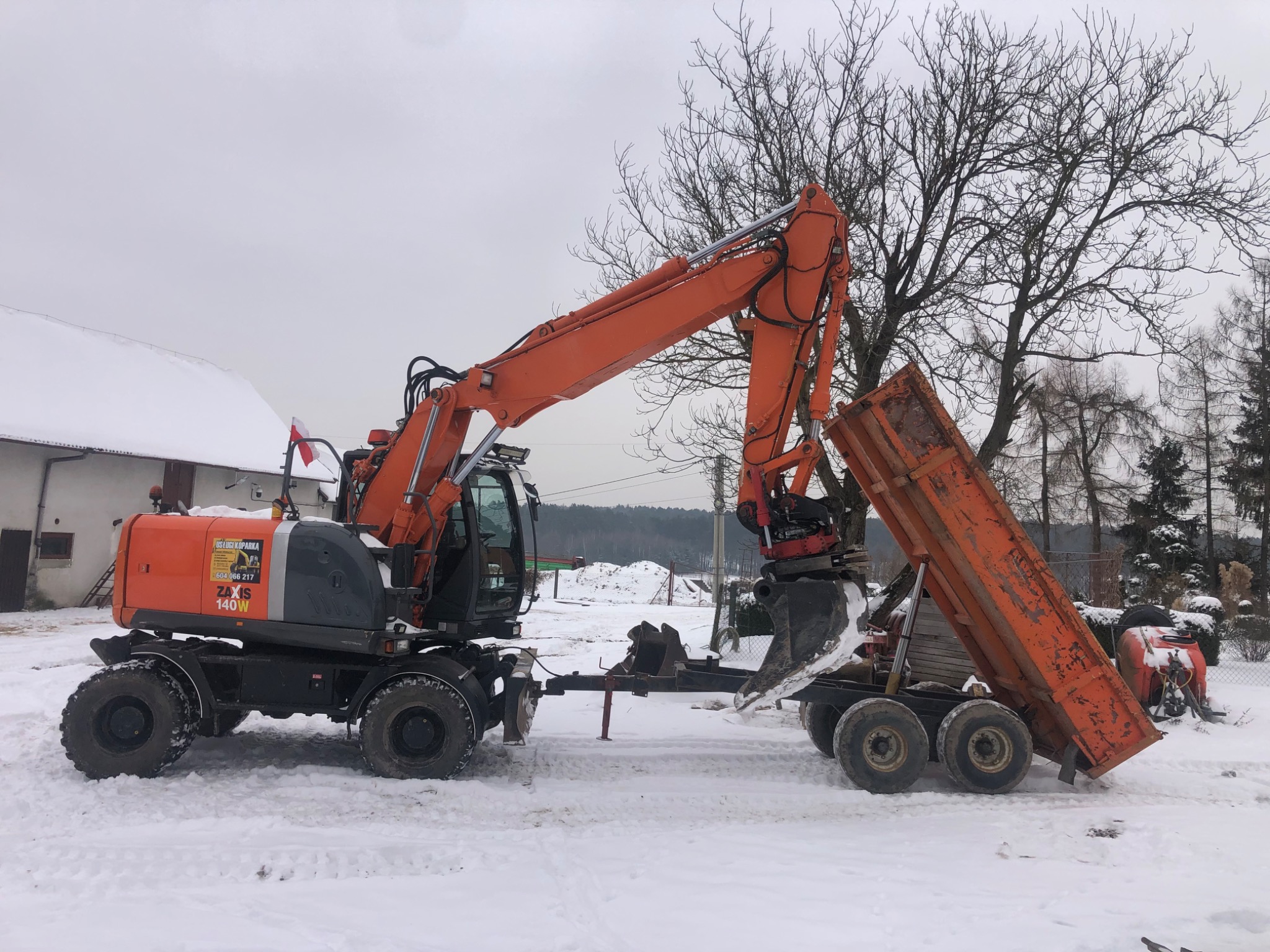 Pomarańczowa koparka kołowa Zaxis 140W na śniegu, z łyżką nad wywrotką; w tle budynek z polską flagą, drzewa i zaspy śnieżne.