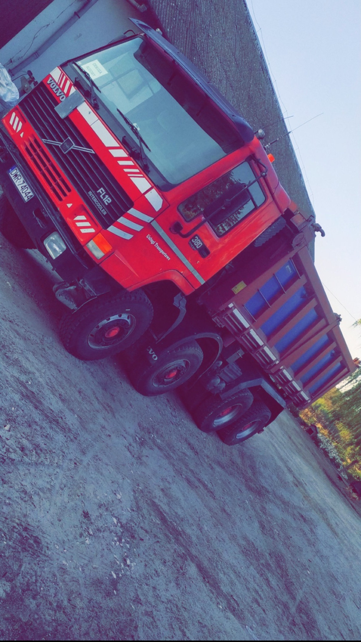 Czerwona wywrotka Volvo FL12 z niebieską skrzynią ładunkową, zaparkowana na szarej, utwardzonej powierzchni, widok z przodu i boku, z widocznym numerem rejestracyjnym DWR 74044.