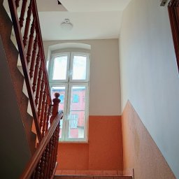 Dawid Wójcik - Klatka schodowa w budynku wielorodzinnym z pomarańczową lamperią i drewnianą balustradą, widok z dołu na okno z widokiem na inny budynek.