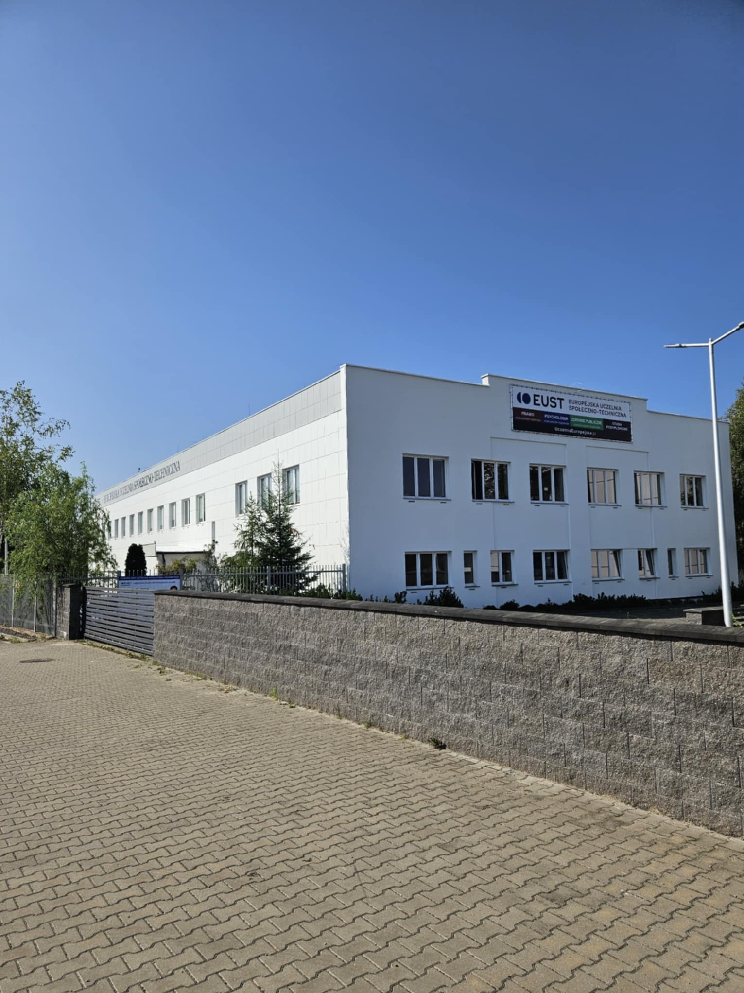 Biały budynek uczelni z widocznym banerem reklamowym, sfotografowany z poziomu chodnika w słoneczny dzień.
