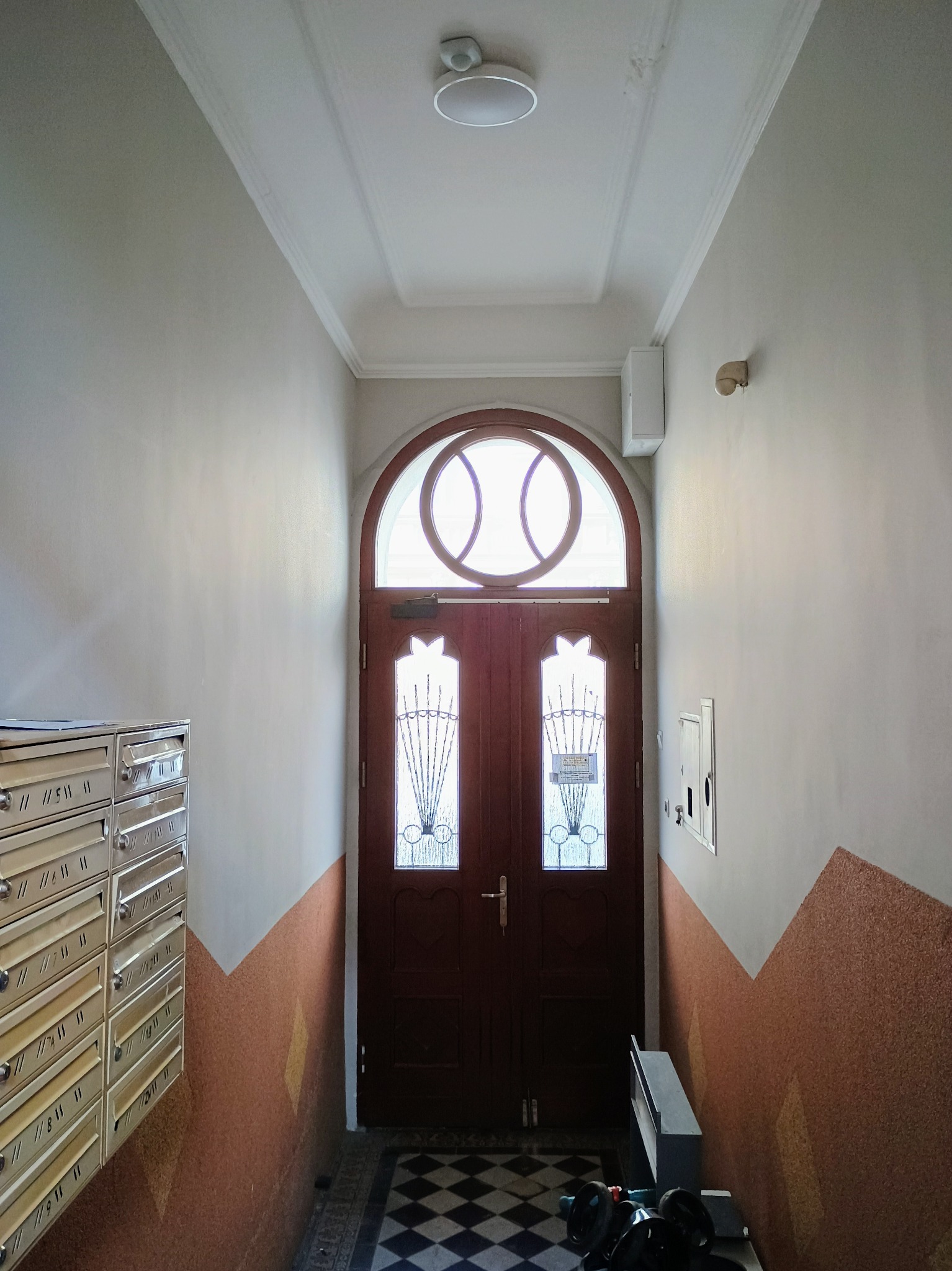 Korytarz w kamienicy z odnowionymi ścianami w kolorze złamanej bieli i pomarańczowym pasem w dolnej części, drewniane drzwi wejściowe z okrągłym oknem na górze i skrzynki pocztowe po lewej stronie.