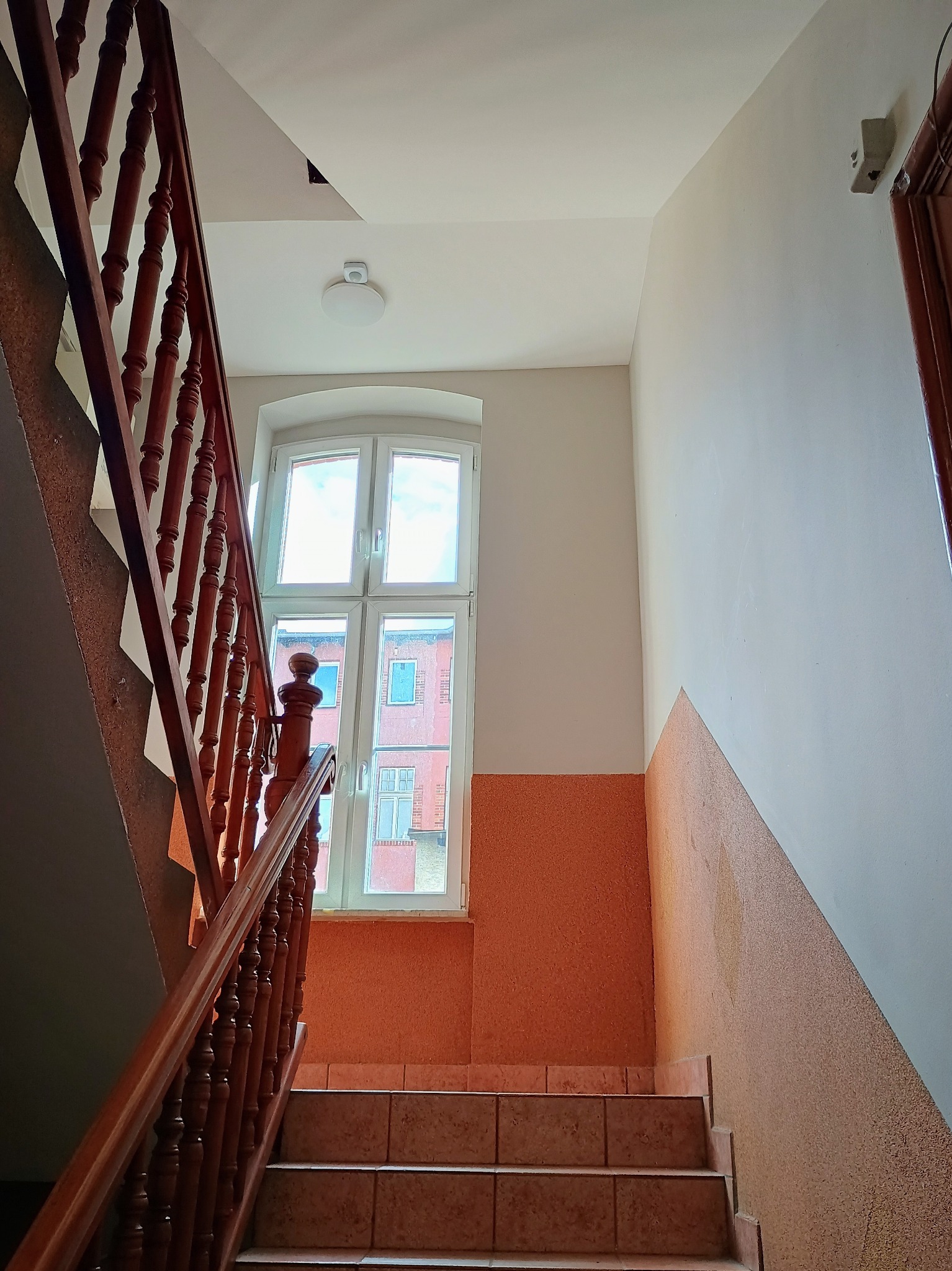 Klatka schodowa w budynku wielorodzinnym z pomarańczową lamperią i drewnianą balustradą, widok z dołu na okno z widokiem na inny budynek.