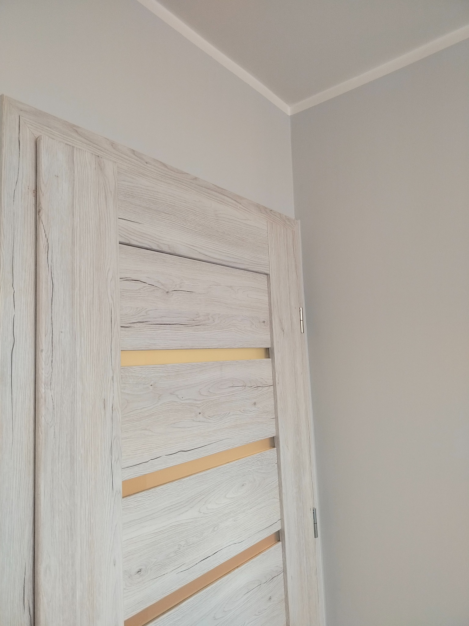 Zamknięte, wewnętrzne drzwi z okleiną imitującą jasne drewno z poziomymi, beżowymi intarsjami na tle szarej ściany i białego sufitowego panelu.