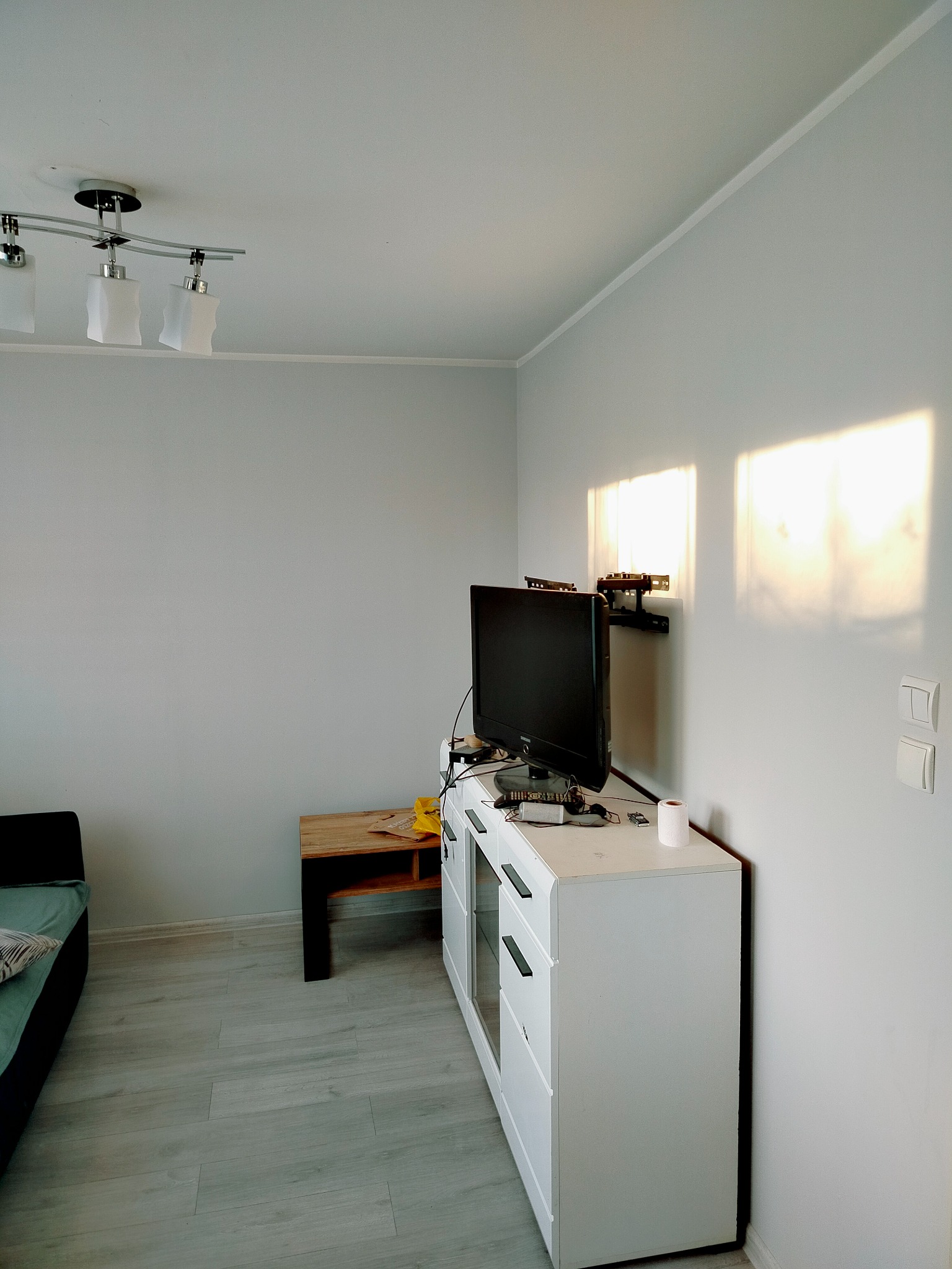 Jasne wnętrze pokoju po odświeżeniu ścian, z widocznym telewizorem na komodzie, drewnianym stolikiem i fragmentem sofy. Ściany pomalowane na jasny odcień szarości, z promieniami słonecznymi...