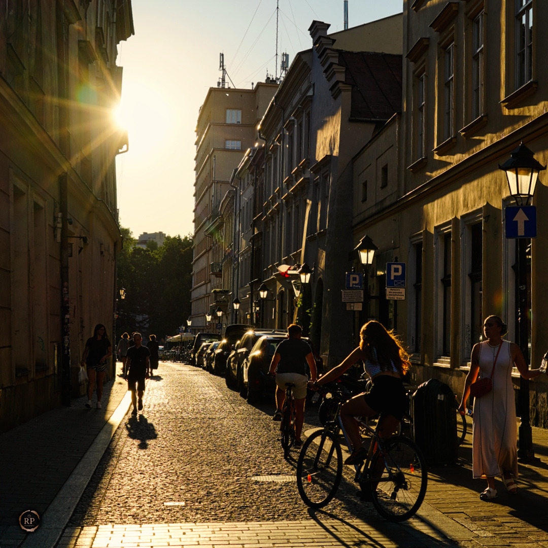 Ulica w blasku zachodzącego słońca: rowerzyści, piesi, zaparkowane auta, stylowe latarnie i architektoniczne detale kamienic tworzą malowniczy miejski krajobraz.