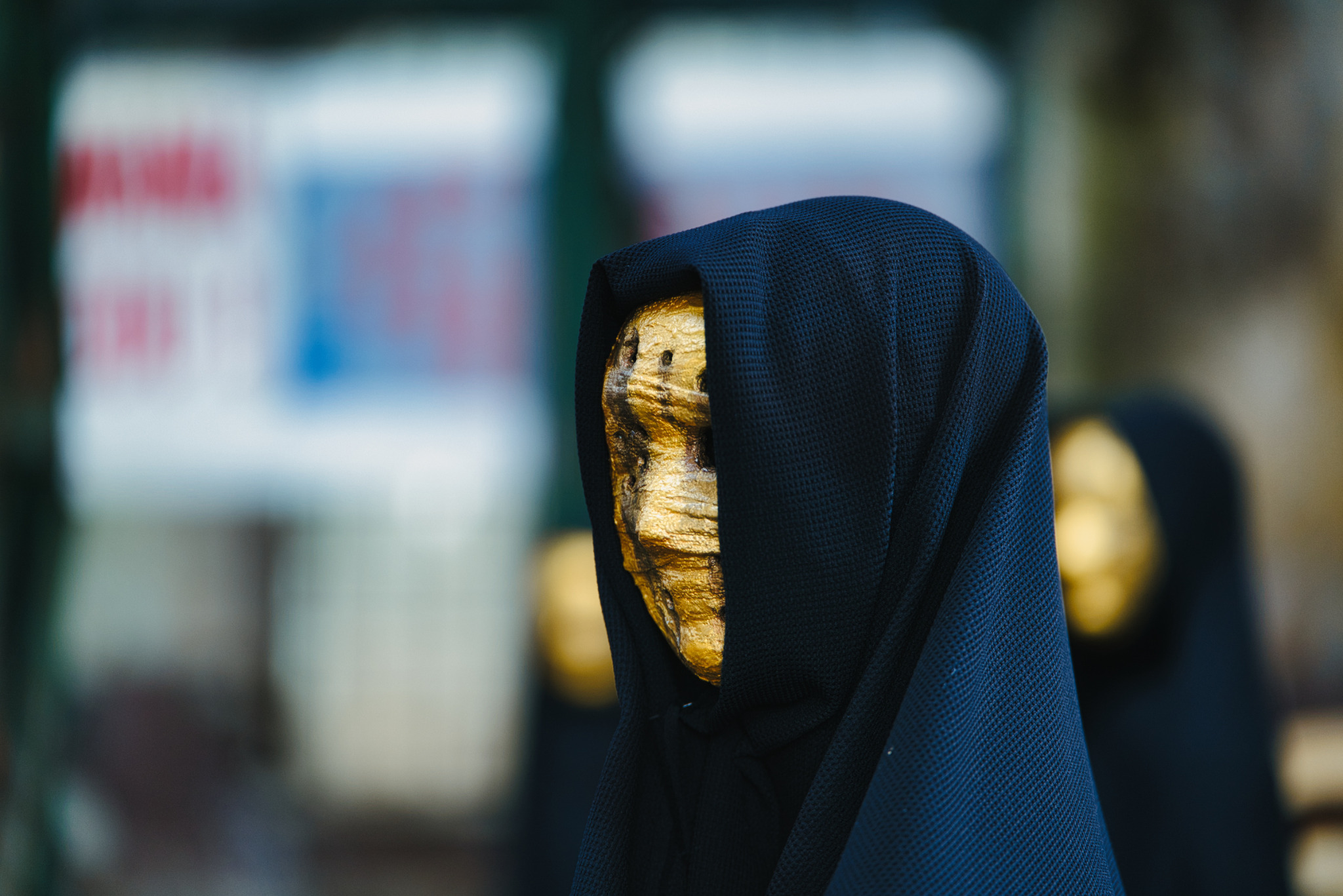 Złota maska o chropowatej fakturze, zakryta czarnym, teksturowanym kapturem na rozmytym tle. Ujęcie z bliska, centralne.
