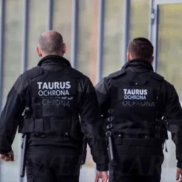 Dwóch ochroniarzy w czarnych uniformach z napisem 'TAURUS Ochrona' na plecach, idących wzdłuż przeszklonej ściany.