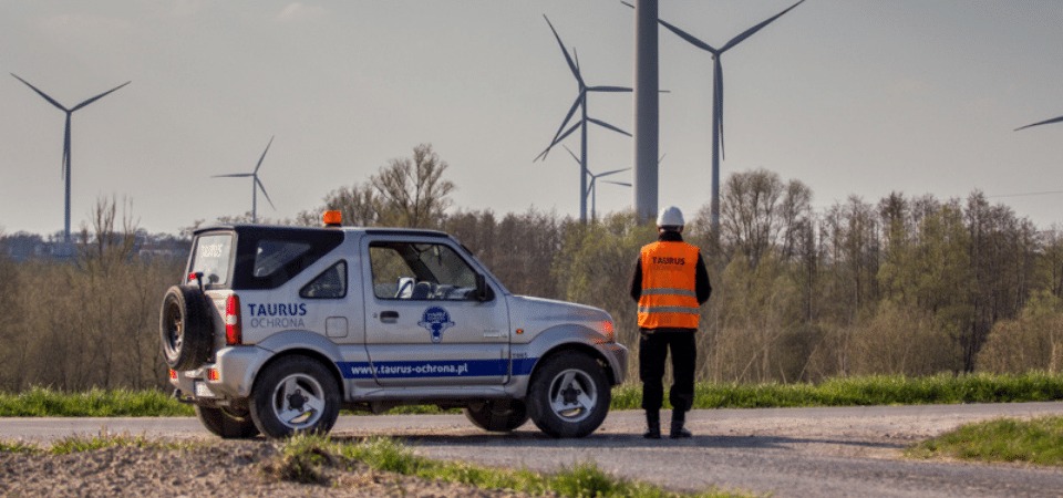 Pracownik ochrony w pomarańczowej kamizelce i białym kasku stoi obok srebrnego samochodu terenowego z logo firmy ochroniarskiej Taurus na tle farmy wiatrowej.