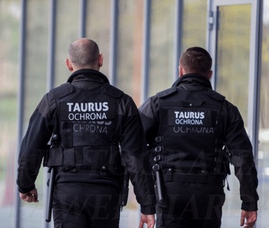 Dwóch ochroniarzy w czarnych uniformach z napisem 'TAURUS Ochrona' na plecach, idących wzdłuż przeszklonej ściany.