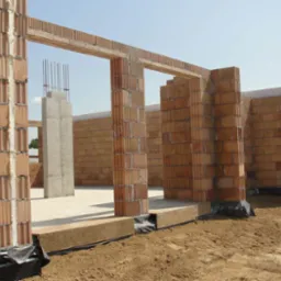 Wznoszenie ścian z czerwonej cegły dziurawki na fundamencie z widoczną izolacją, słup żelbetowy ze zbrojeniem w tle, stan surowy otwarty.