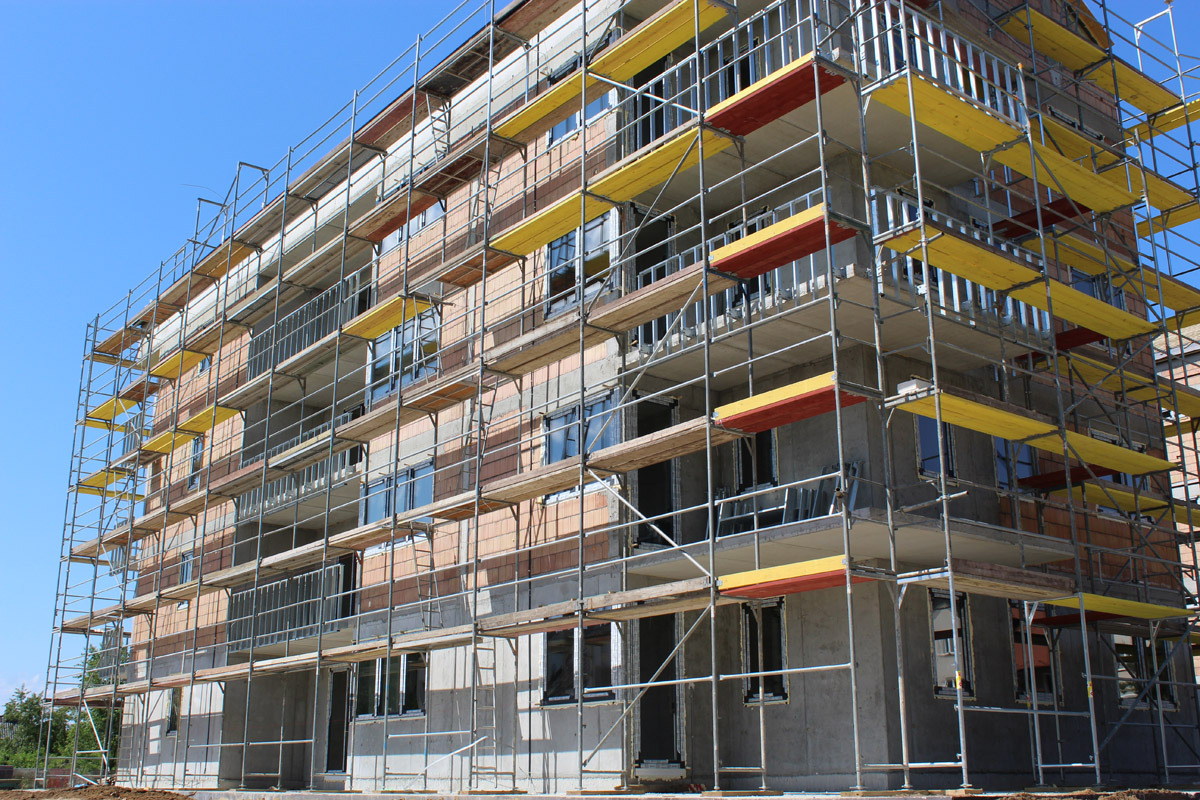 Wielopiętrowy budynek w trakcie wznoszenia, z widocznymi szarymi ścianami z betonu komórkowego, częściowo obłożonymi czerwoną cegłą, otoczony metalowymi rusztowaniami z żółtymi i czerwonymi deskami...