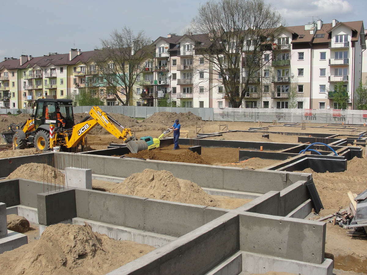 Wylane betonowe ławy fundamentowe na placu budowy, z żółtą koparko-ładowarką JCB i pracownikiem zagęszczającym grunt w tle, na tle wielorodzinnych budynków mieszkalnych.