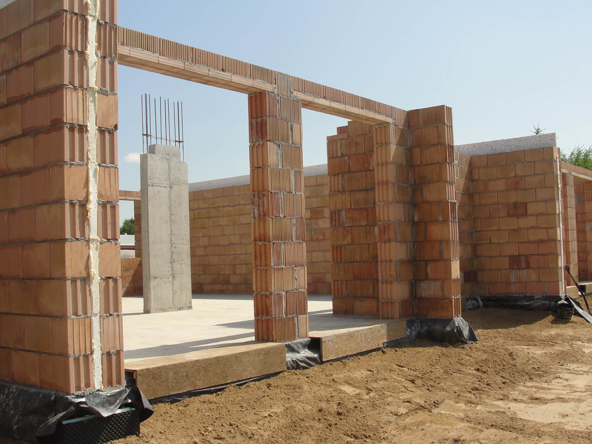 Wznoszenie ścian z czerwonej cegły dziurawki na fundamencie z widoczną izolacją, słup żelbetowy ze zbrojeniem w tle, stan surowy otwarty.