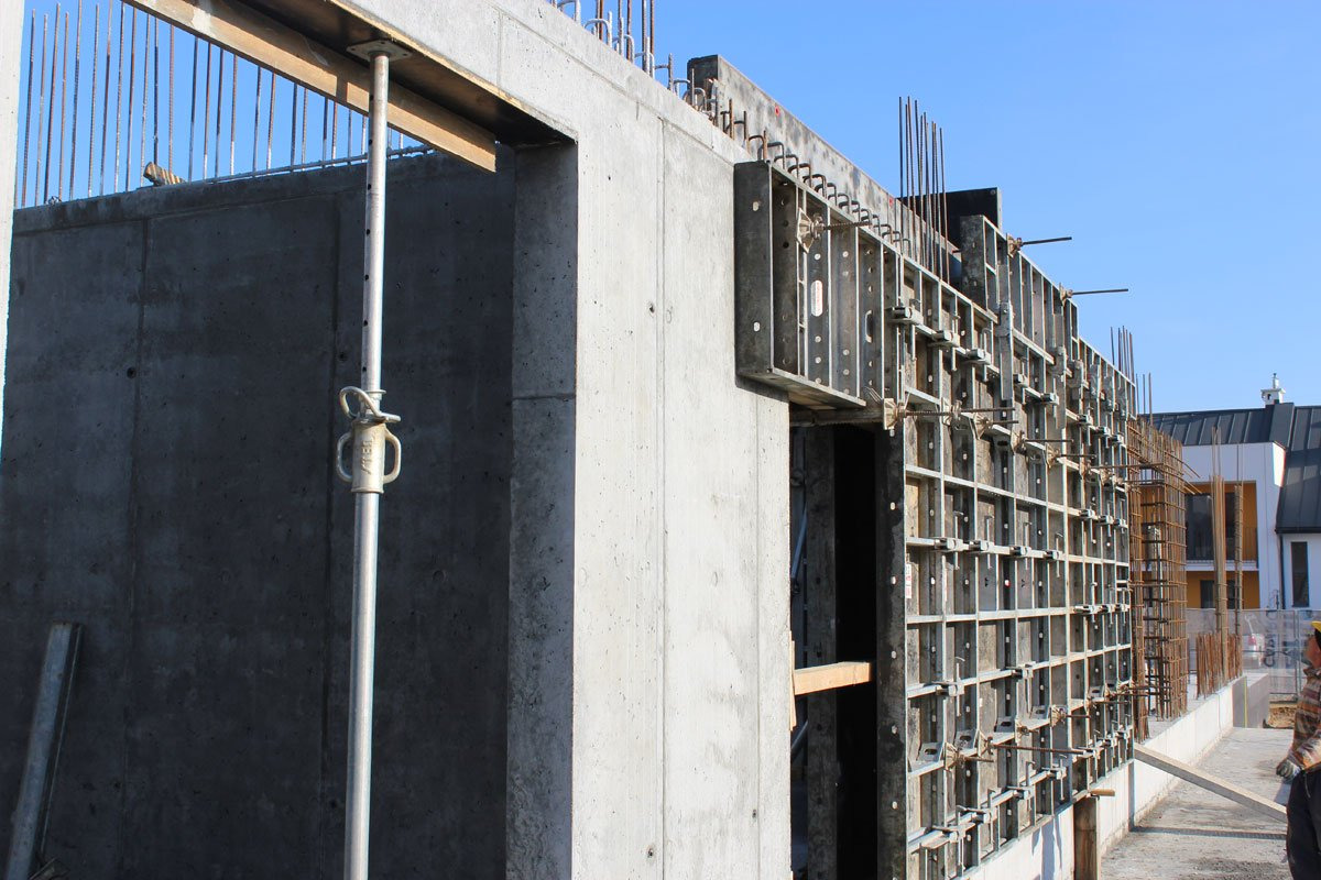 Surowa ściana żelbetowa w trakcie budowy, widoczne szalunki i wystające pręty zbrojeniowe, wsporniki oraz fragment budynku w tle.