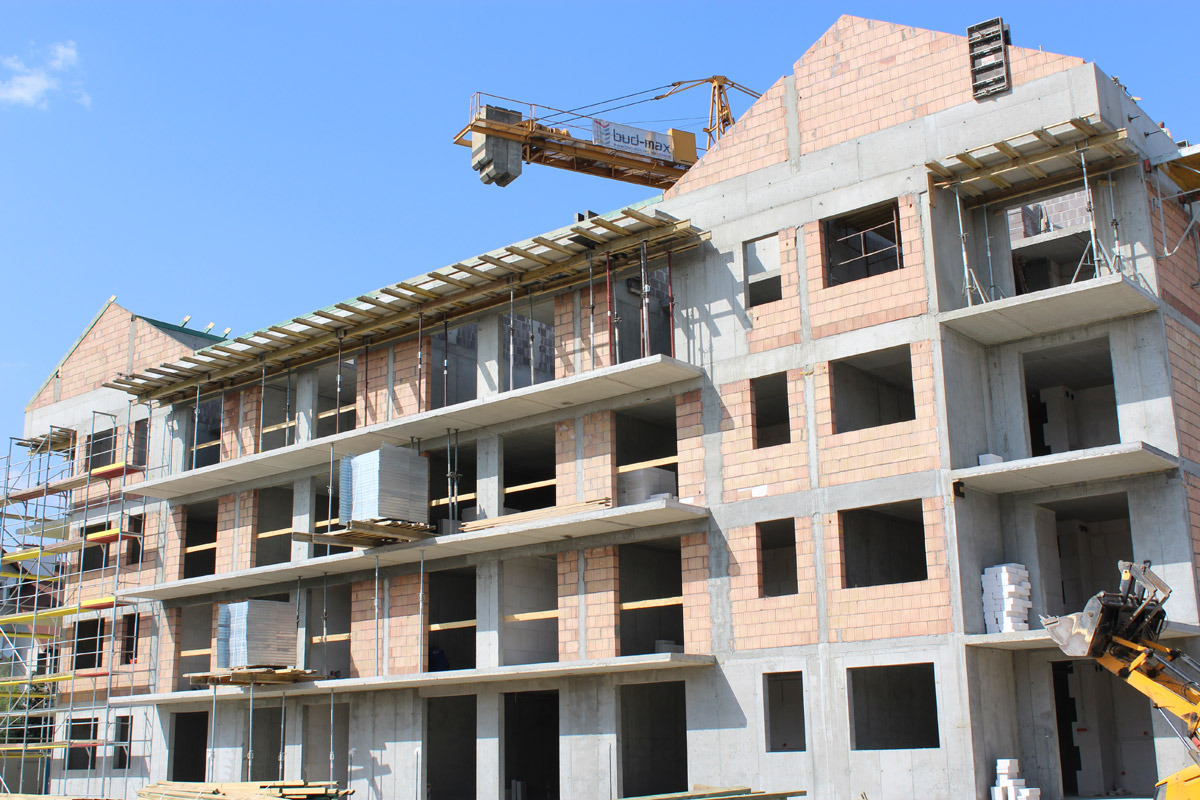 Wielopiętrowy budynek w trakcie wznoszenia z betonu i cegły, widoczny żuraw budowlany na tle błękitnego nieba.