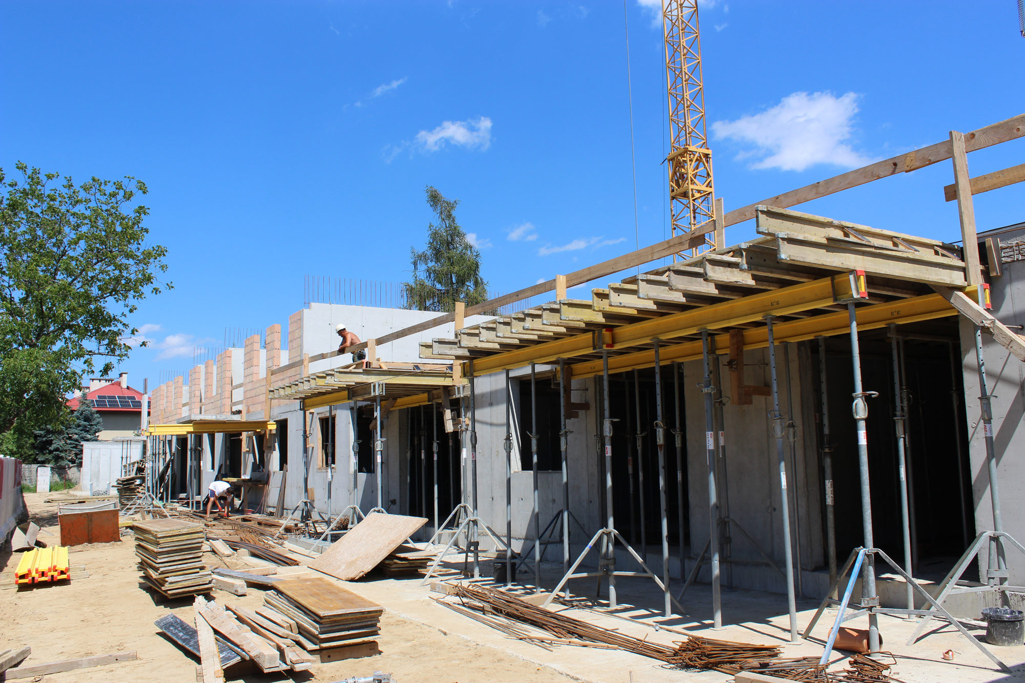 Wznoszenie budynku w stanie surowym otwartym: szalunki na stropie, widoczni pracownicy w trakcie prac, dźwig w tle, elementy zbrojenia i stosy płyt szalunkowych na placu budowy.