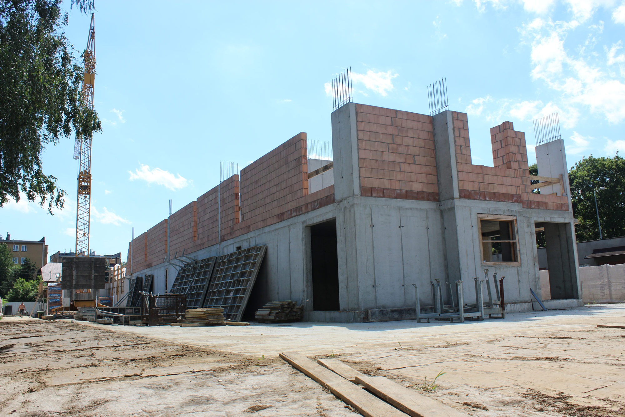 Surowy stan budynku w trakcie budowy: betonowy szkielet z fragmentami murowanych ścian z czerwonej cegły, widoczne zbrojenia, szalunki i dźwig w tle, teren budowy z piaskiem i deskami.