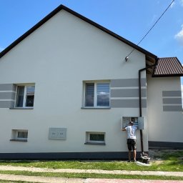 MD BUD - Dwukondygnacyjny dom z ocieploną i pomalowaną elewacją w odcieniach bieli i szarości. Mężczyzna w białej koszulce sprawdza skrzynkę gazową na ścianie. Widoczne okna z roletami i brązowy dach.