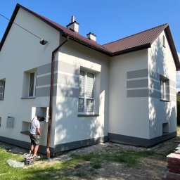 MD BUD - Pracownik maluje elewację domu jednorodzinnego na biało z szarymi pasami, widoczne narzędzia malarskie i wiadro z farbą, brązowy dach i rynny, błękitne niebo w tle.