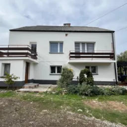 Dwukondygnacyjny dom z białymi ścianami i ciemnymi balkonami, widok z poziomu gruntu na front budynku, szare wykończenie cokołu, zieleń w ogrodzie.