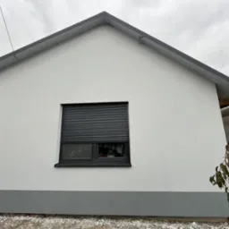 Świeżo otynkowana elewacja domu jednorodzinnego w odcieniach szarości i bieli, z widocznym oknem z roletą zewnętrzną, wykonana w pochmurny dzień.