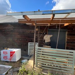 MD BUD - Prace wykończeniowe przy drewnianym domu z zadaszeniem: montaż rolet zewnętrznych, widoczne paczki styropianu NTB i częściowo zbudowany płot.