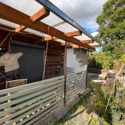 MD BUD - Pracownik w beżowej bluzie montuje roletę zewnętrzną na drewnianej ścianie budynku z widoczną konstrukcją zadaszenia z poliwęglanu i ogrodzeniem z metalowych sztachet.