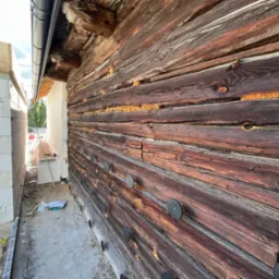 Fragment drewnianej ściany domu z widocznymi szczelinami wypełnionymi pianką izolacyjną i zamocowanymi okrągłymi elementami mocującymi, sąsiadujący z fragmentem ściany z białych bloczków.