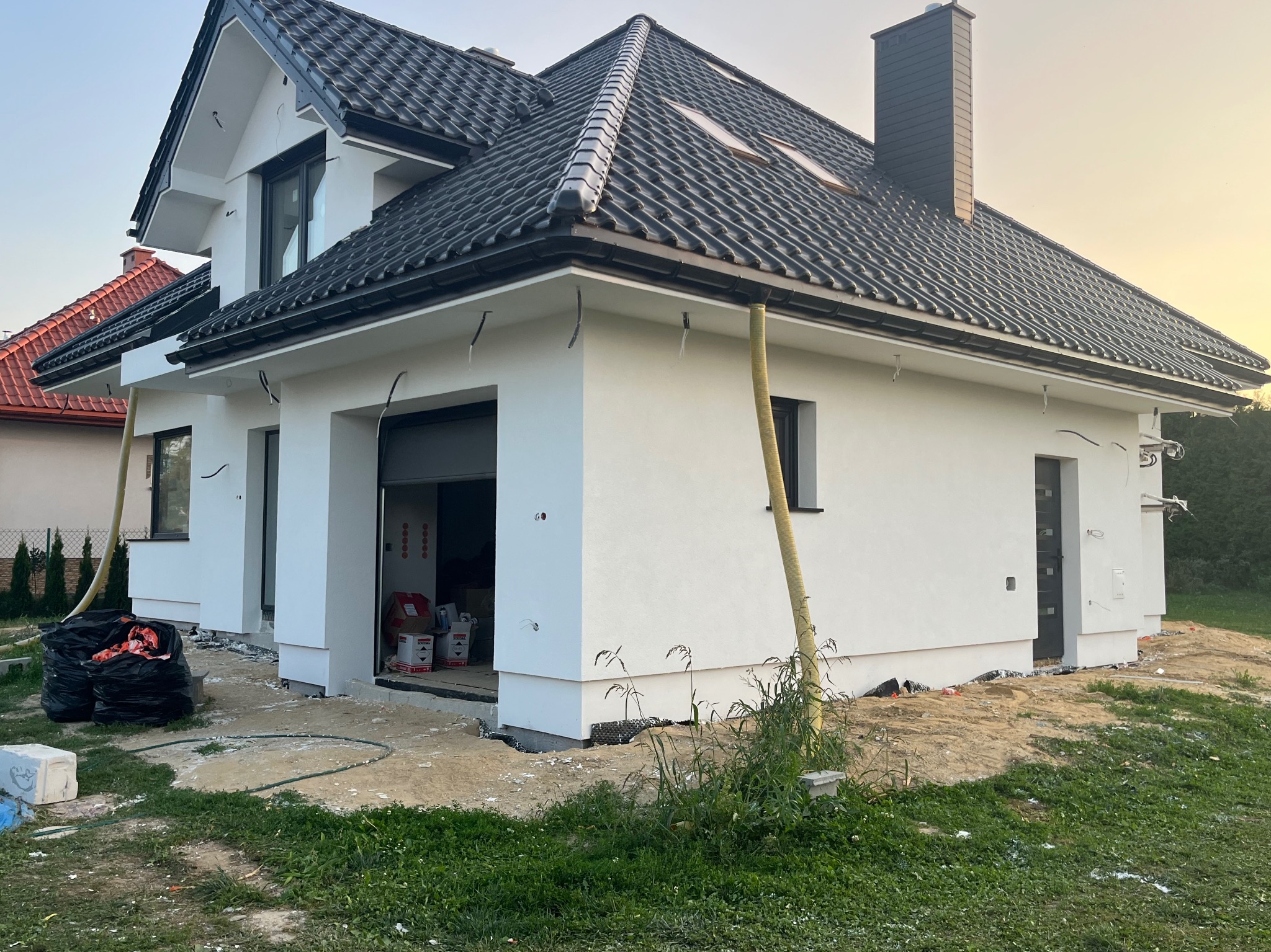 Niedokończony dom jednorodzinny z białą elewacją, czarną dachówką i widocznymi elementami instalacji na zewnątrz, worki z odpadami budowlanymi przy ścianie, teren wokół w trakcie prac wykończeniowych.