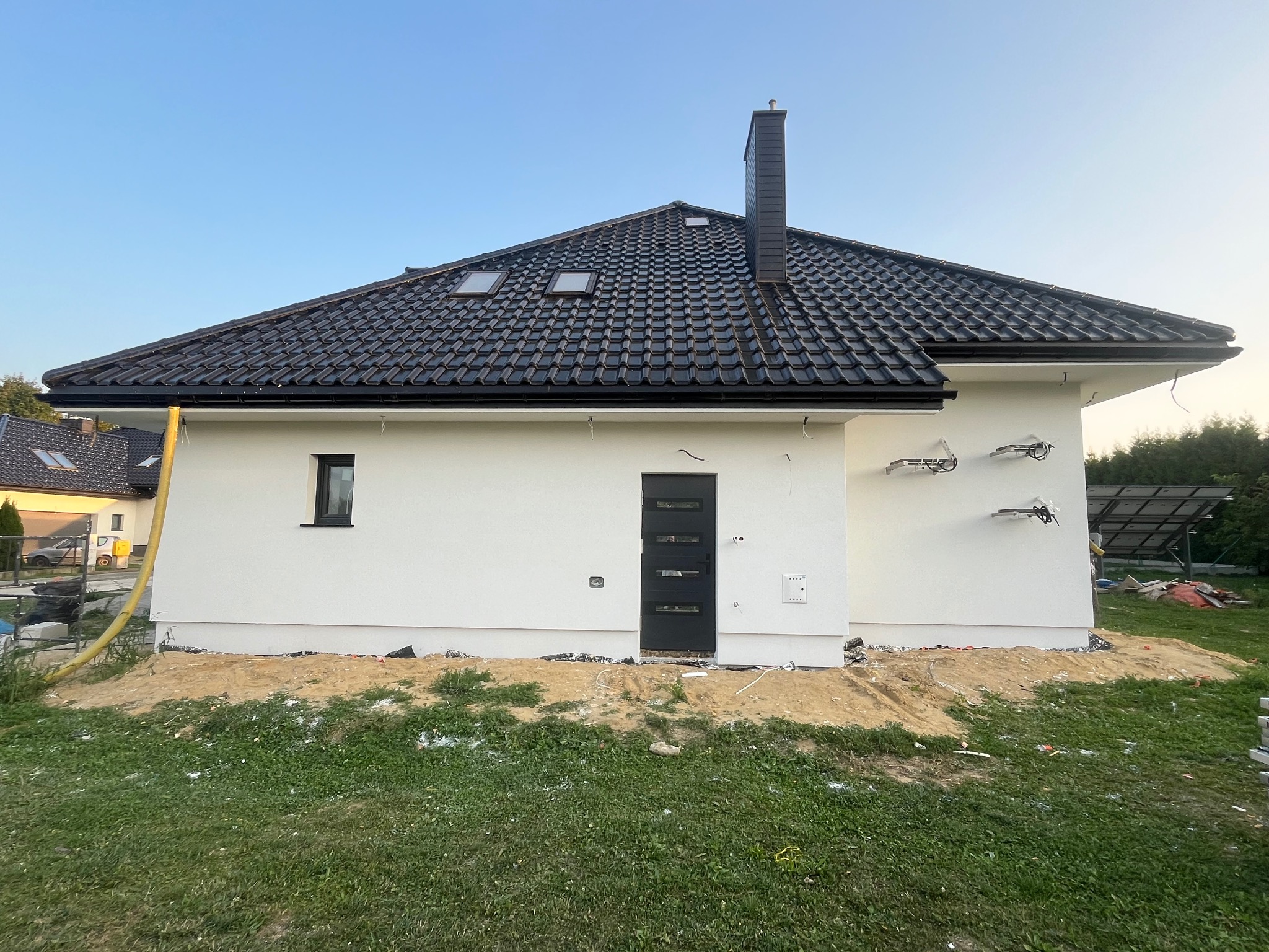 Nowoczesny dom jednorodzinny z białą elewacją, czarnym dachem z dachówki, oknami dachowymi i czarnymi elementami wykończeniowymi, widoczne przygotowania do prac wykończeniowych wokół budynku.