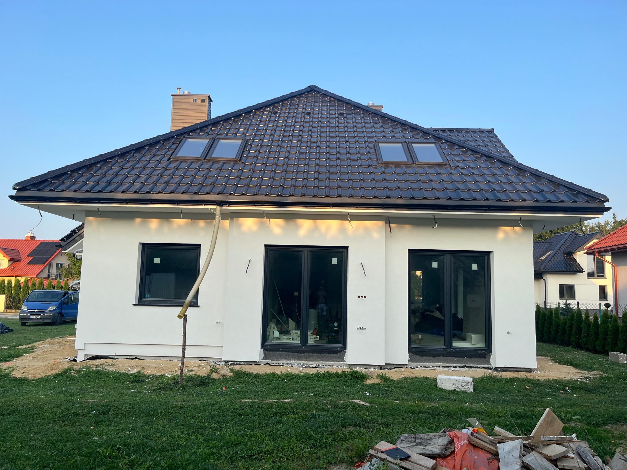 Nowoczesny dom jednorodzinny z białą elewacją, czarną dachówką, oknami dachowymi i czarną stolarką okienną; teren wokół domu częściowo zagospodarowany, widoczne ślady prac budowlanych.